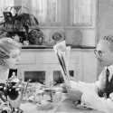 A picture of a man and woman sitting at a dinner table circa 1950. The man is reading a newspaper while the women listens.