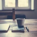 Image of a desk with a laptop, coffee cup, notebook, and pen